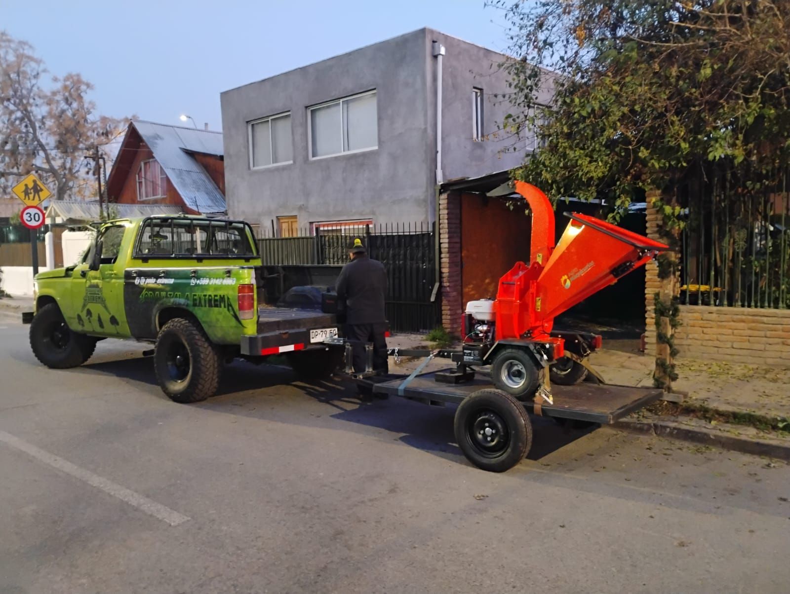 Un camión verde con remolque transporta una trituradora de madera roja. Una persona está cerca de la trituradora en la calle.