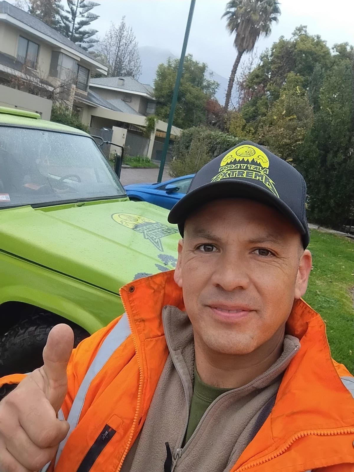 Un hombre con una chaqueta naranja hace un gesto de aprobación con el pulgar delante de un coche verde.