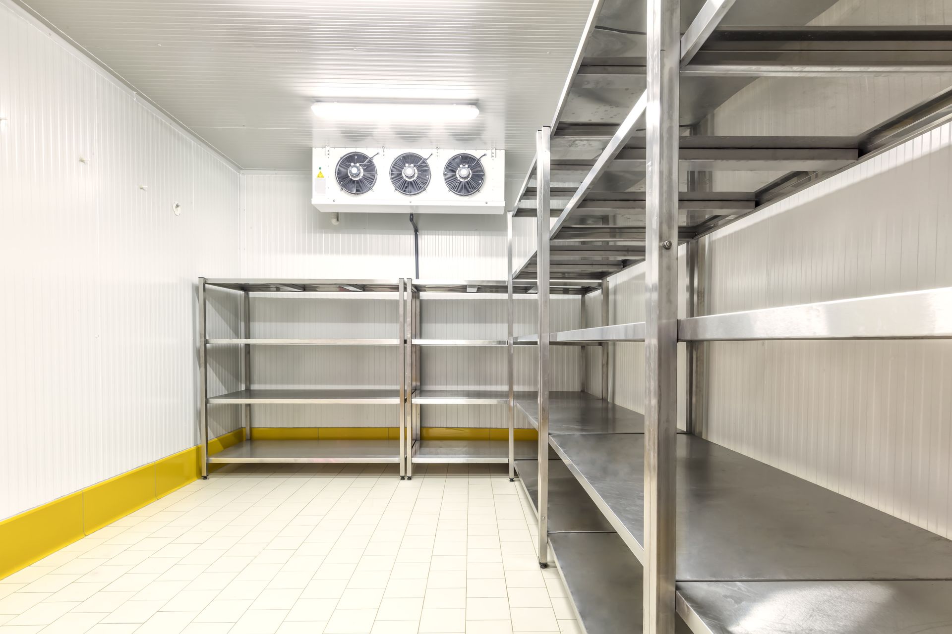Empty walk-in freezer with metal shelving and refrigeration unit. White walls and ceiling, yellow floor trim.