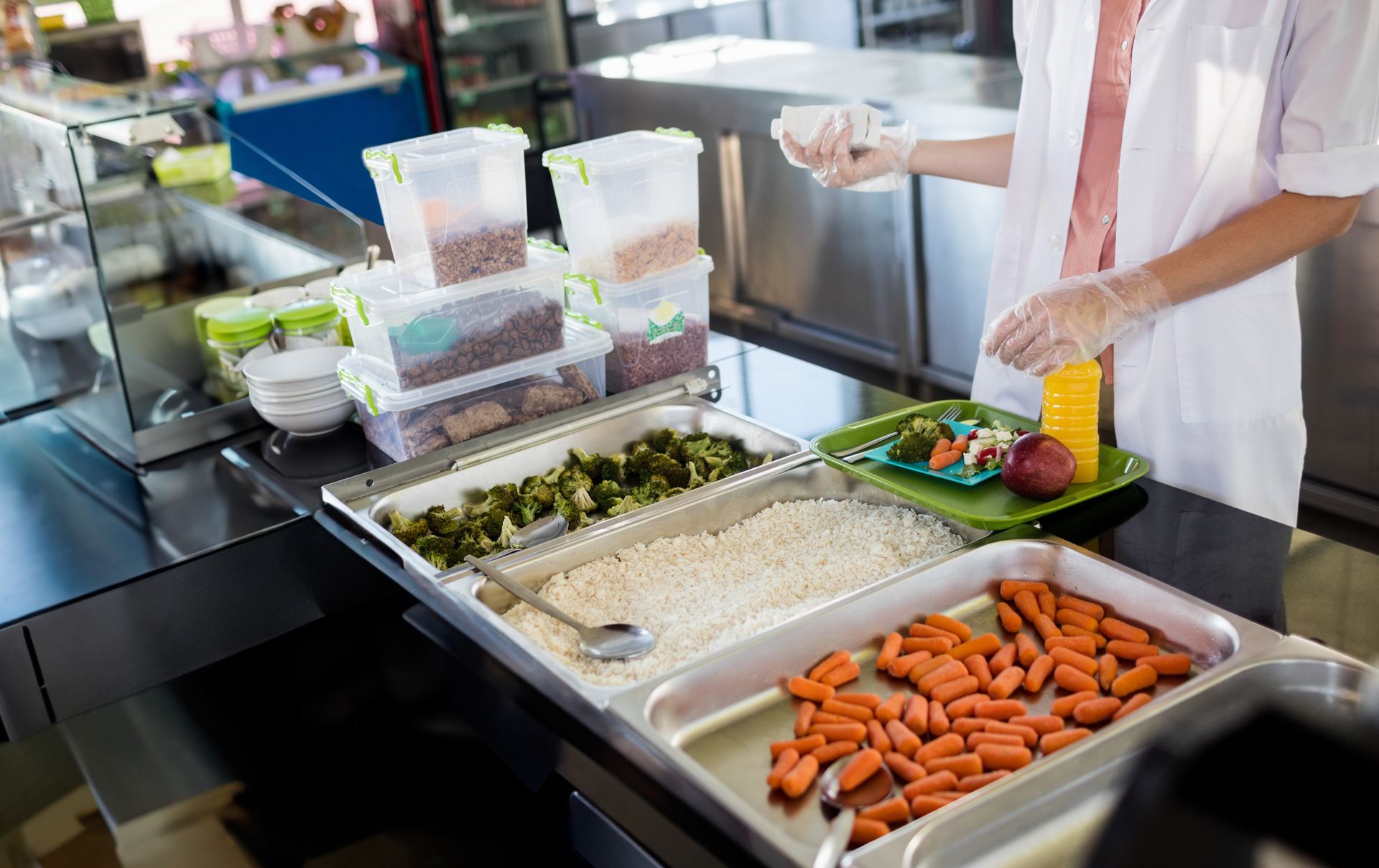 A person in gloves and a white coat serving food from a cafeteria line with vegetables and grains.