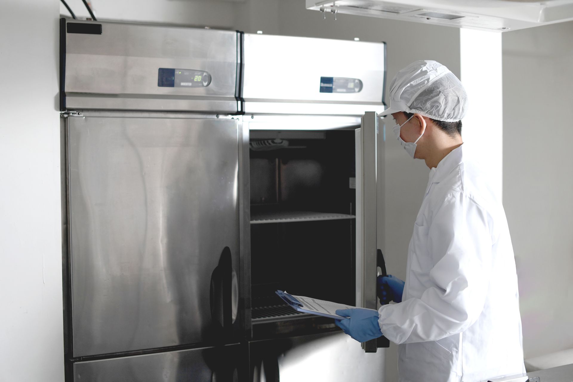 Person in lab coat and hairnet inspecting a stainless steel refrigerator in a lab.