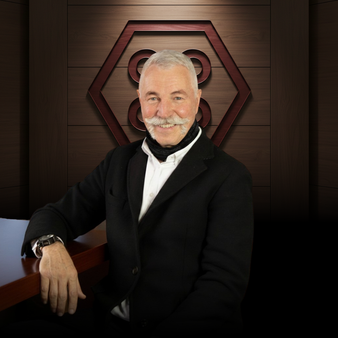 A man in a navy blazer and white shirt poses in front of a wood-paneled wall featuring a hexagonal geometric logo.