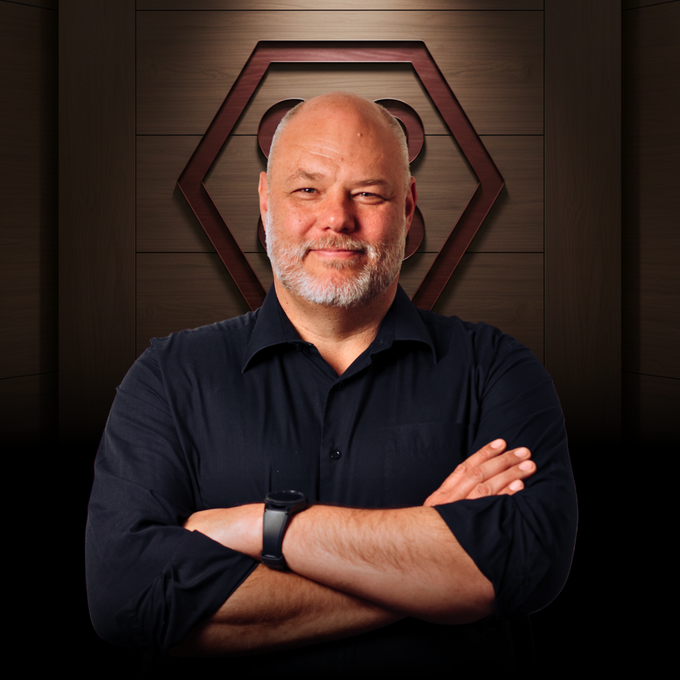 A man in a navy blazer and white shirt poses in front of a wood-paneled wall featuring a hexagonal geometric logo.