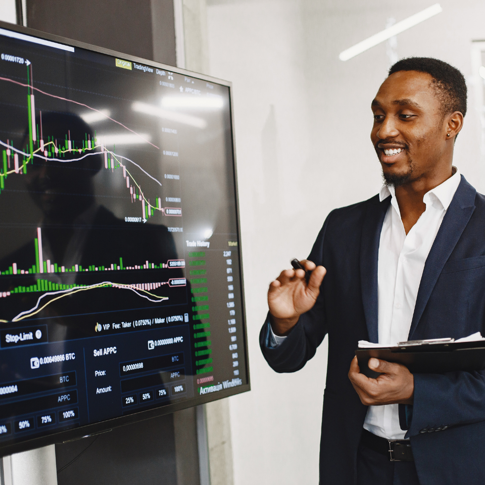 A professional in a suit gestures toward a digital monitor displaying financial stock market data and charts.