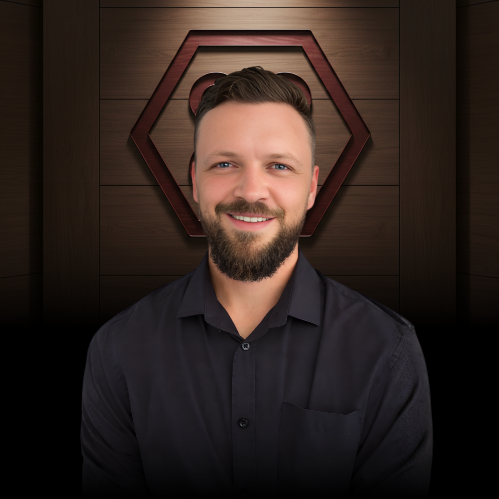 A smiling man with a short, groomed beard wears a black button-down shirt in front of a dark, hexagonal wooden backdrop.