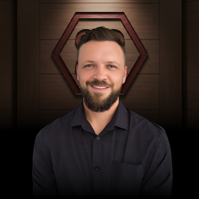 A smiling man with a short, groomed beard wears a black button-down shirt in front of a dark, hexagonal wooden backdrop.