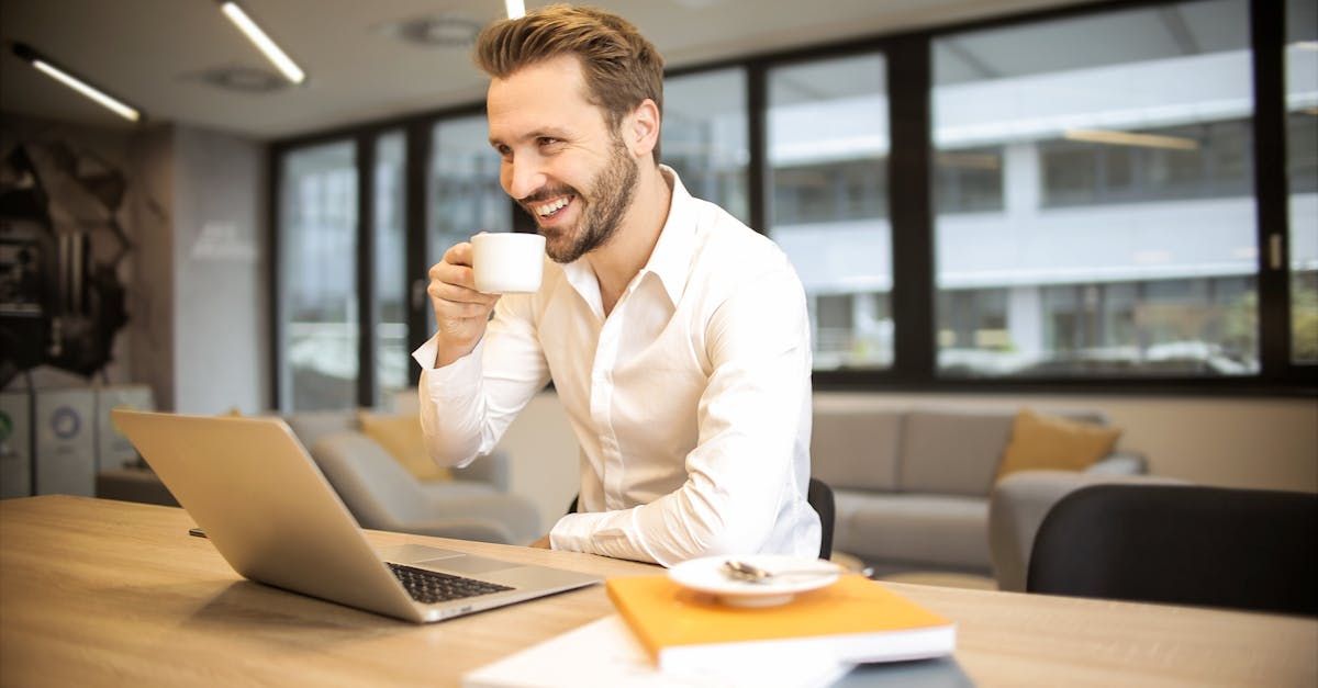 A man is drinking a cup of coffee while using a laptop computer.