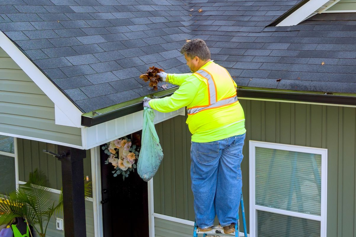 Man on a ladder cleaning a roof gutter, wearing safety vest.