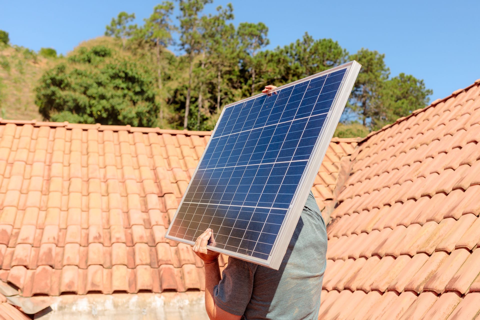 A man is holding a solar panel on top of a tiled roof.