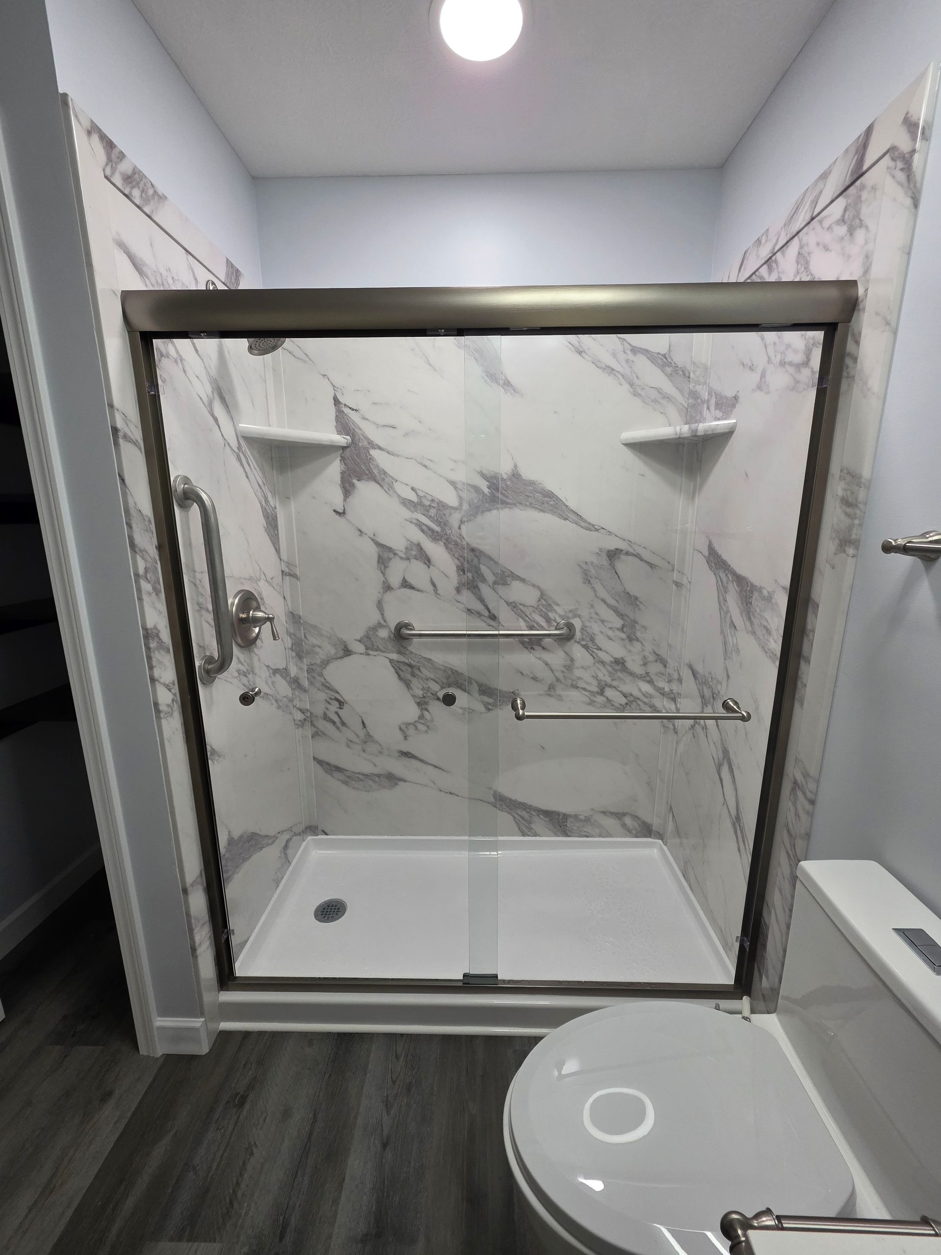 Shower with marble-look walls, sliding glass doors, grab bars, and a white toilet.