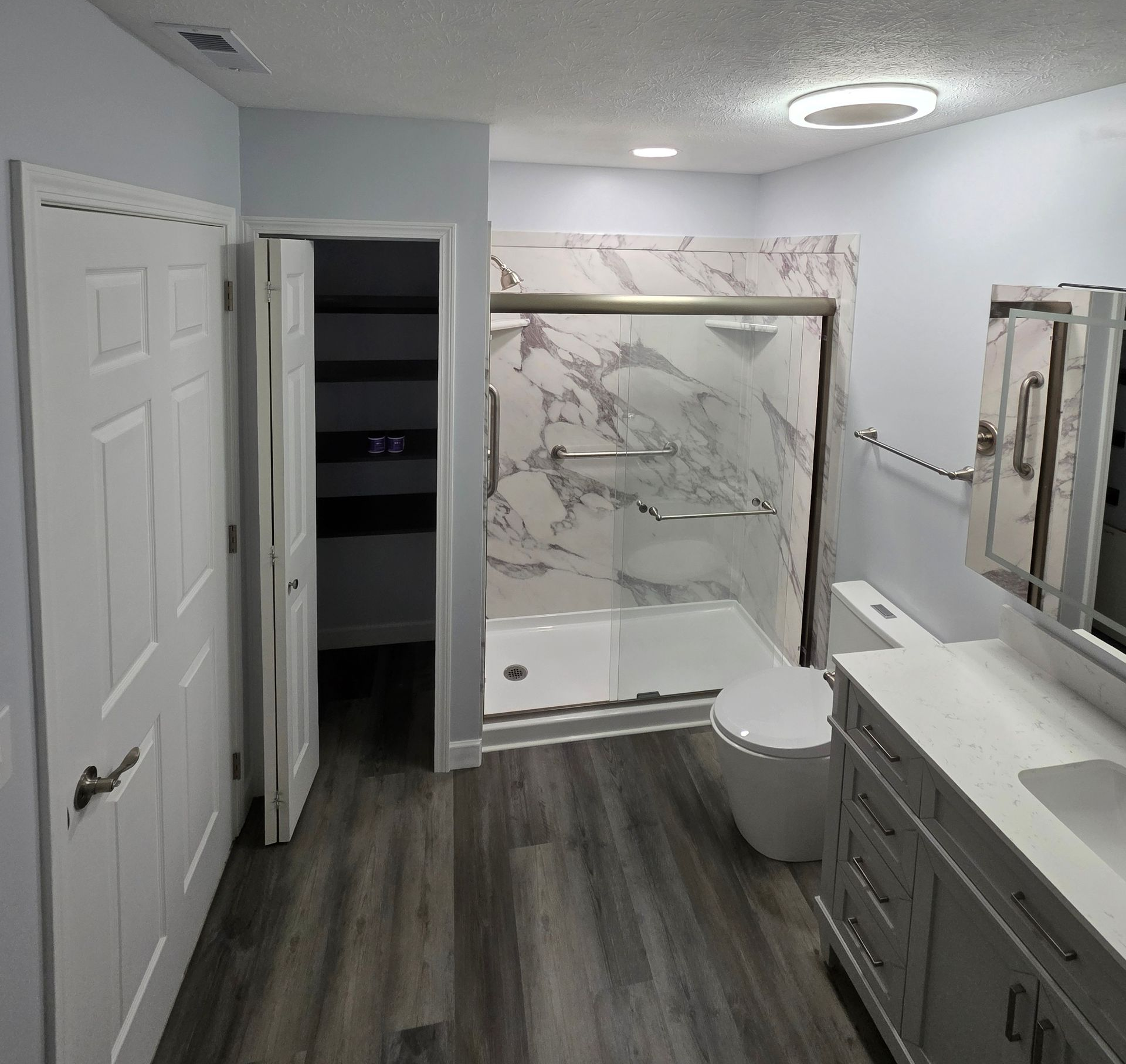 Bathroom with marble shower, gray wood-look floor, light blue walls, white vanity, toilet, and closet door.