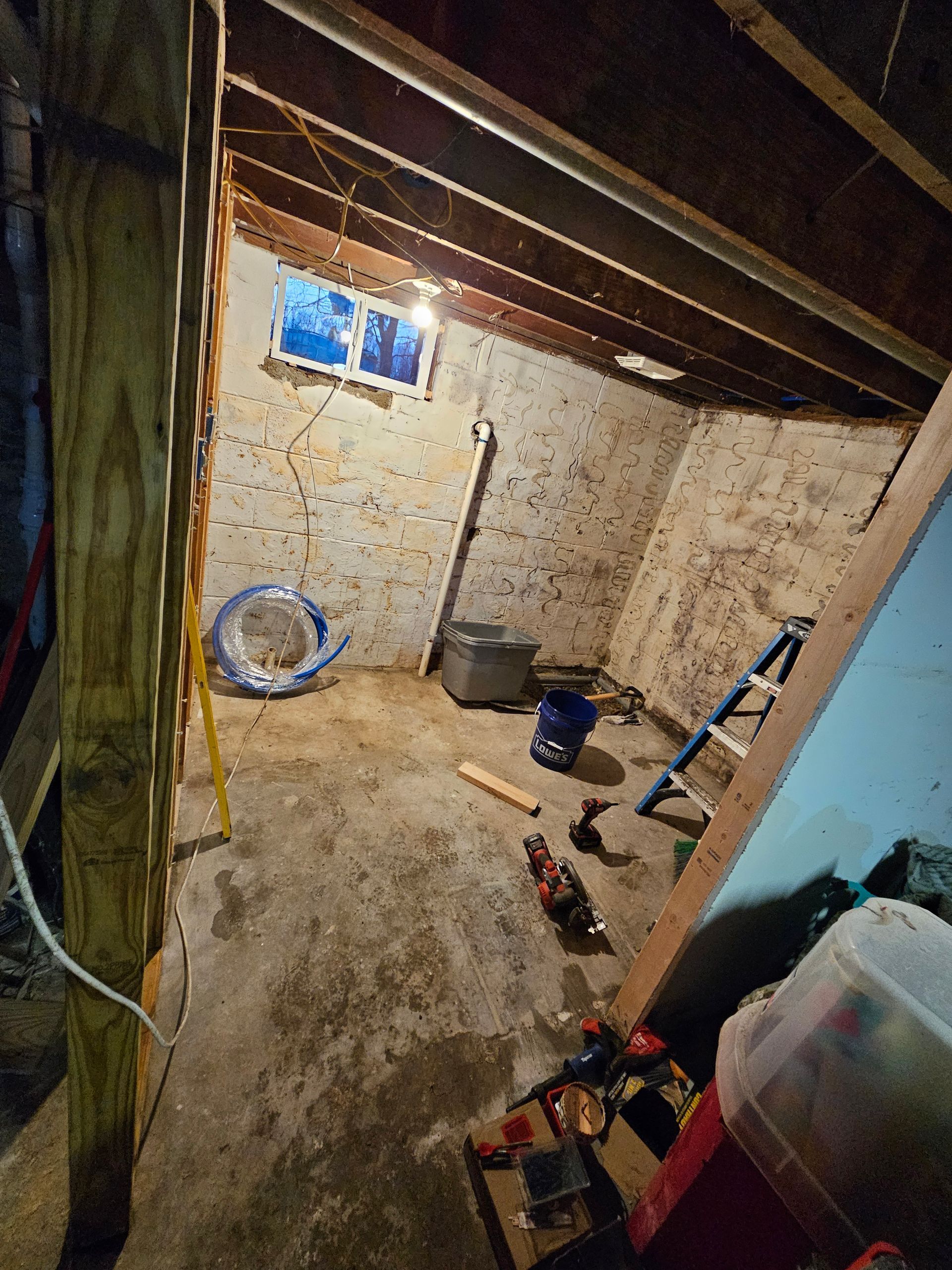 Basement interior with concrete floor, exposed wood beams, small window, and construction materials.