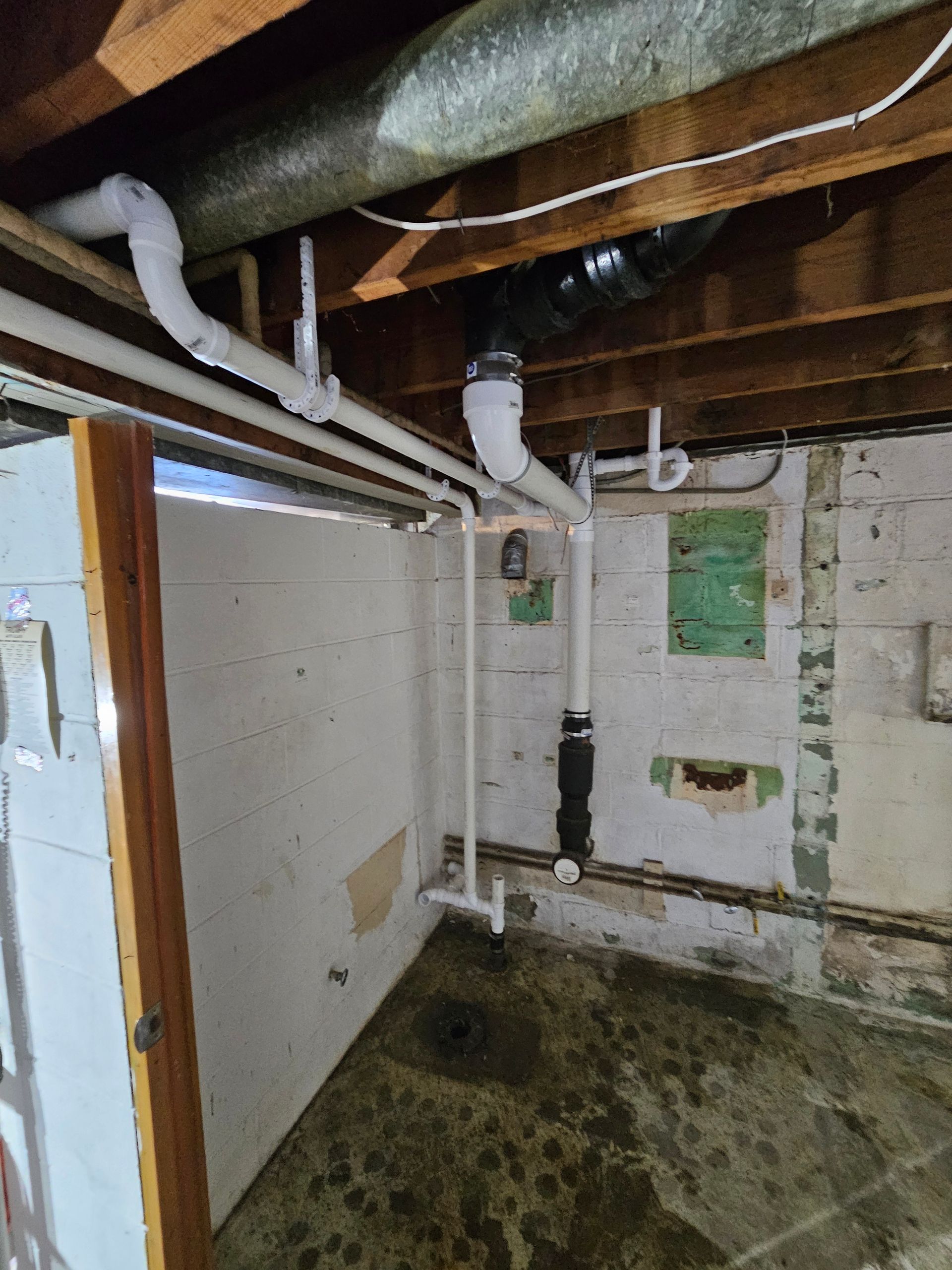 Plumbing pipes in a basement. White pipes run along the ceiling and walls, connected to a drain and vent.