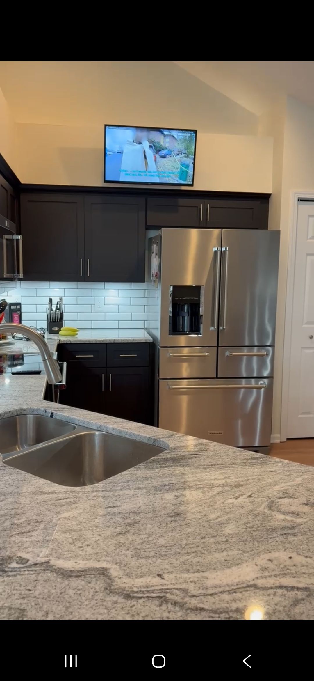 Kitchen with stainless steel refrigerator, dark cabinets, granite countertop, and a TV mounted above.