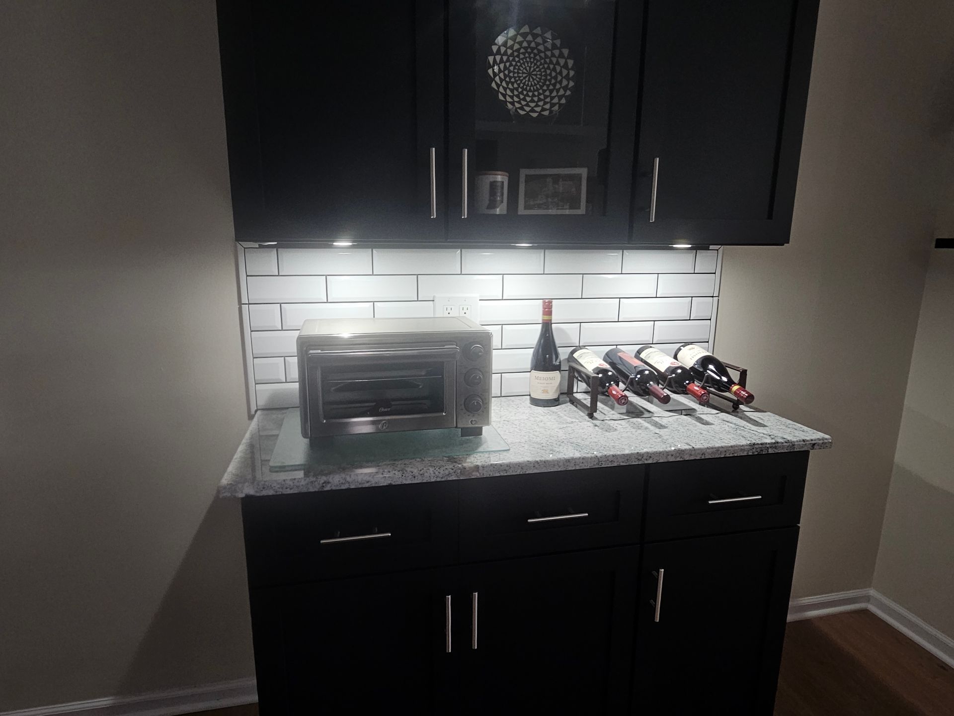 Black cabinets with a countertop, toaster, wine bottles, and under-cabinet lighting.