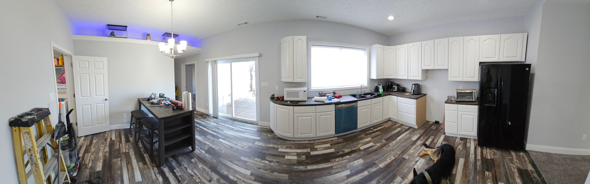 A kitchen with white cabinets, a black refrigerator, and a dark wooden floor. There's a sliding glass door and a chandelier.