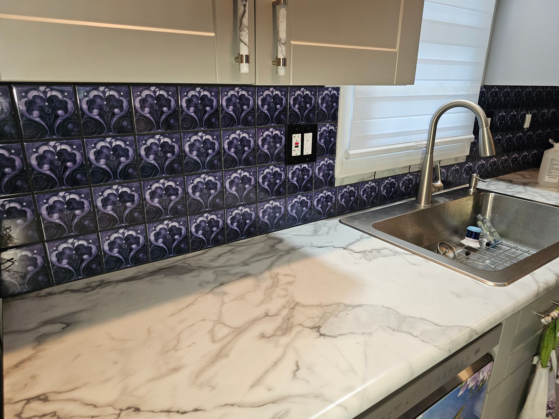 Kitchen counter with marble pattern, dark patterned backsplash, stainless steel sink, and light gray cabinets.