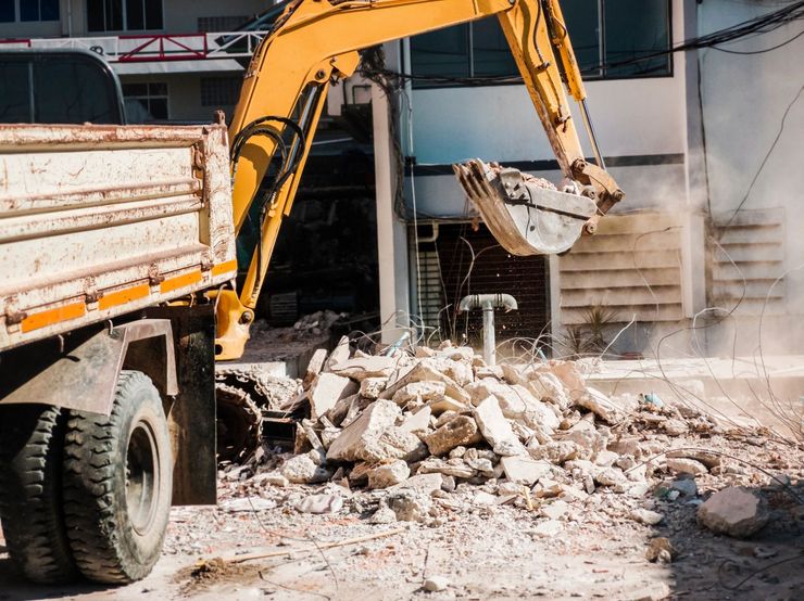 Excavator demolishing rubble beside a truck at a construction site, with dust and broken concrete.