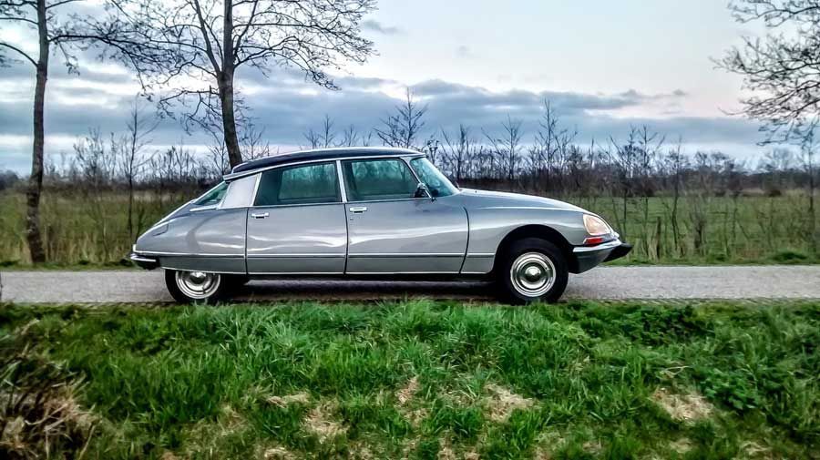 Een zilverkleurige Citroën DS sedan geparkeerd op een weg naast het gras. Avondlucht op de achtergrond.