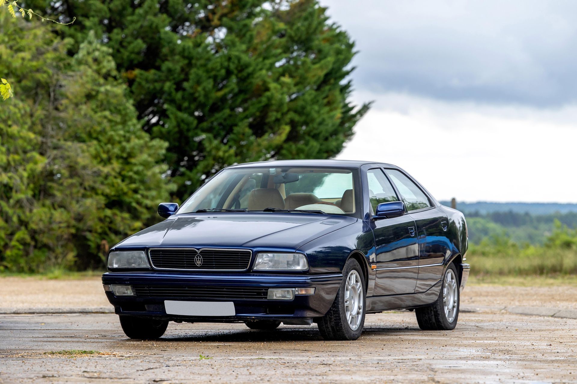 Donkerblauwe Maserati Ghibli coupé geparkeerd op asfalt met een bomenrij op de achtergrond.