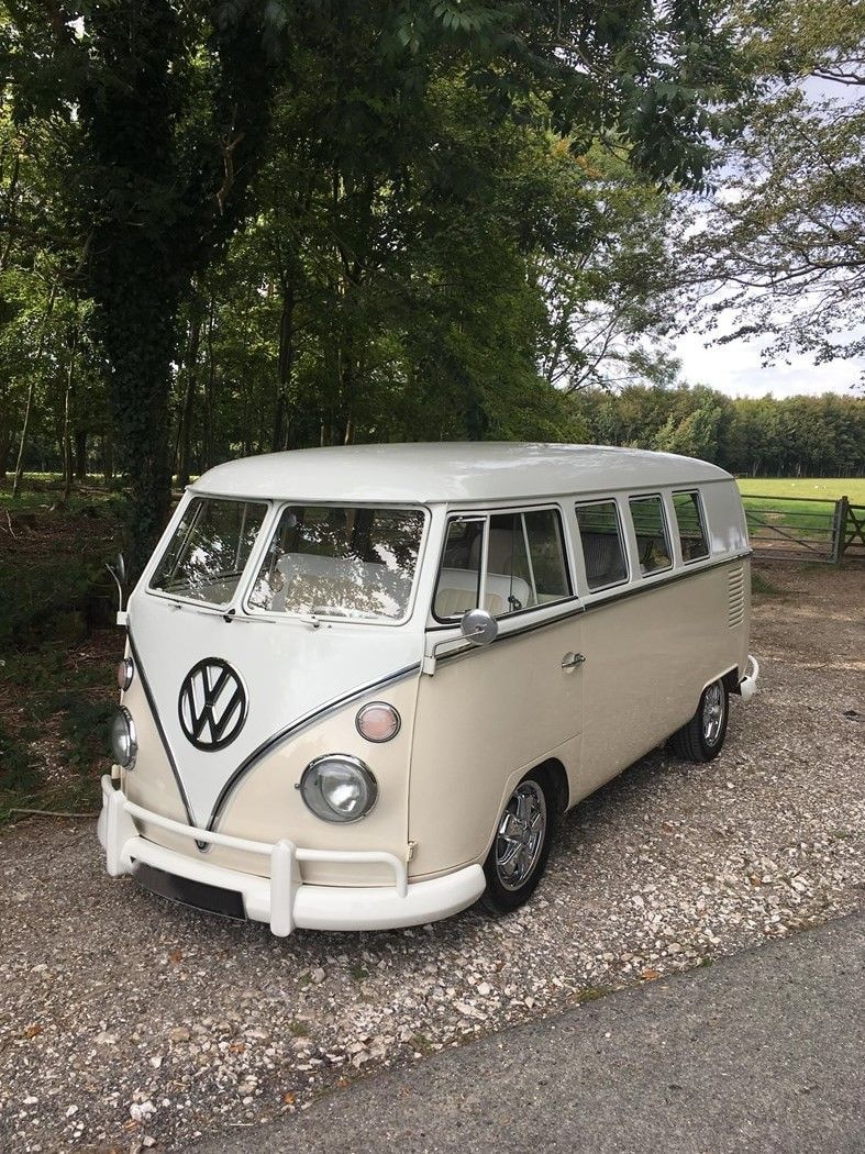 Crème-witte klassieke Volkswagenbus geparkeerd op een grindweg, met bomen op de achtergrond.