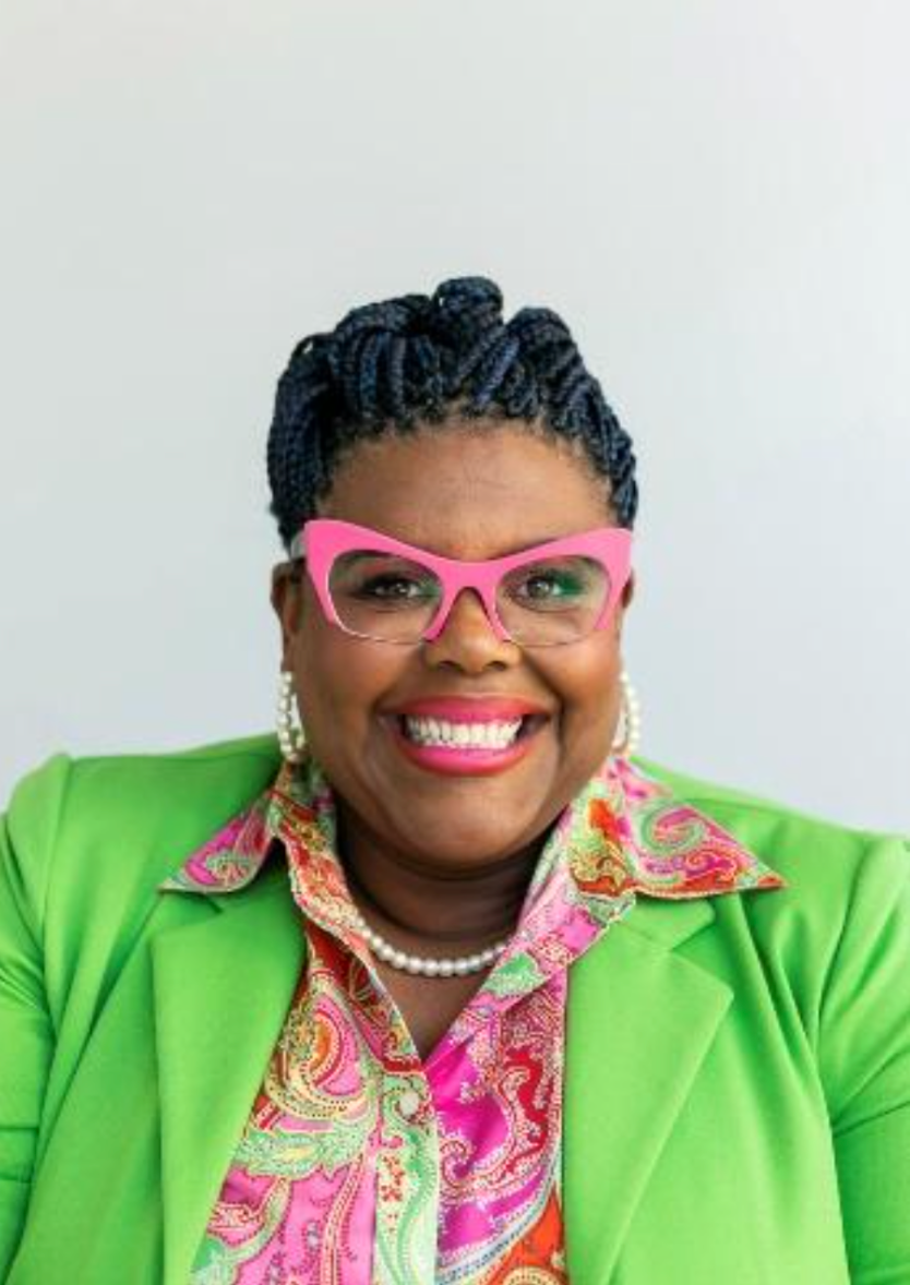 A woman wearing a green jacket and pink glasses is smiling.