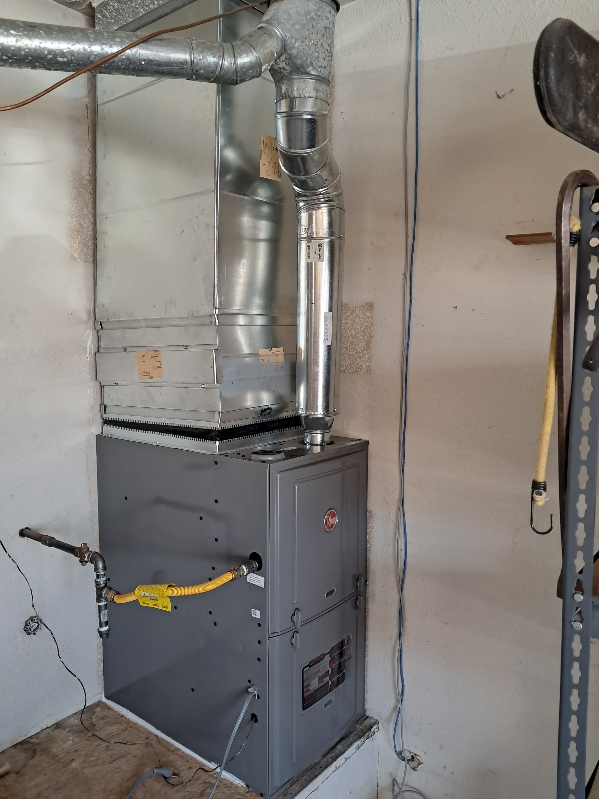 A gray Rheem furnace in a corner, with silver vent pipes and a yellow gas line.