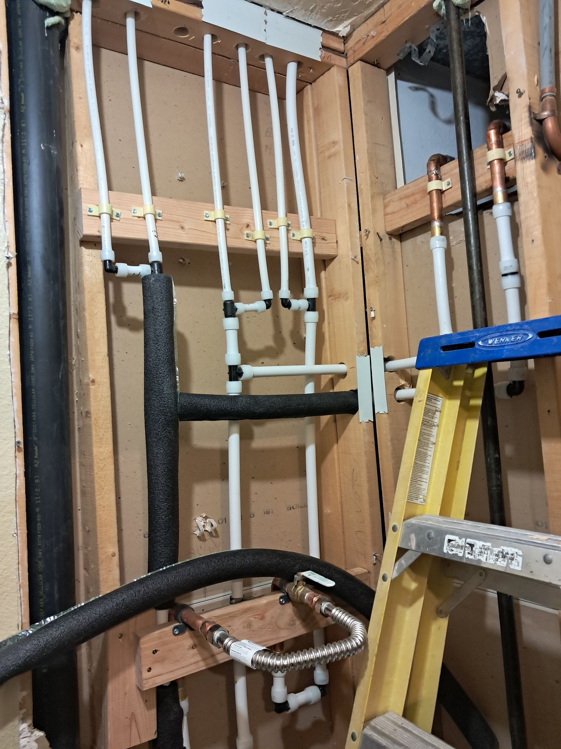 Pipes and plumbing work in a wall cavity, including white, black, and copper pipes, and a yellow ladder.
