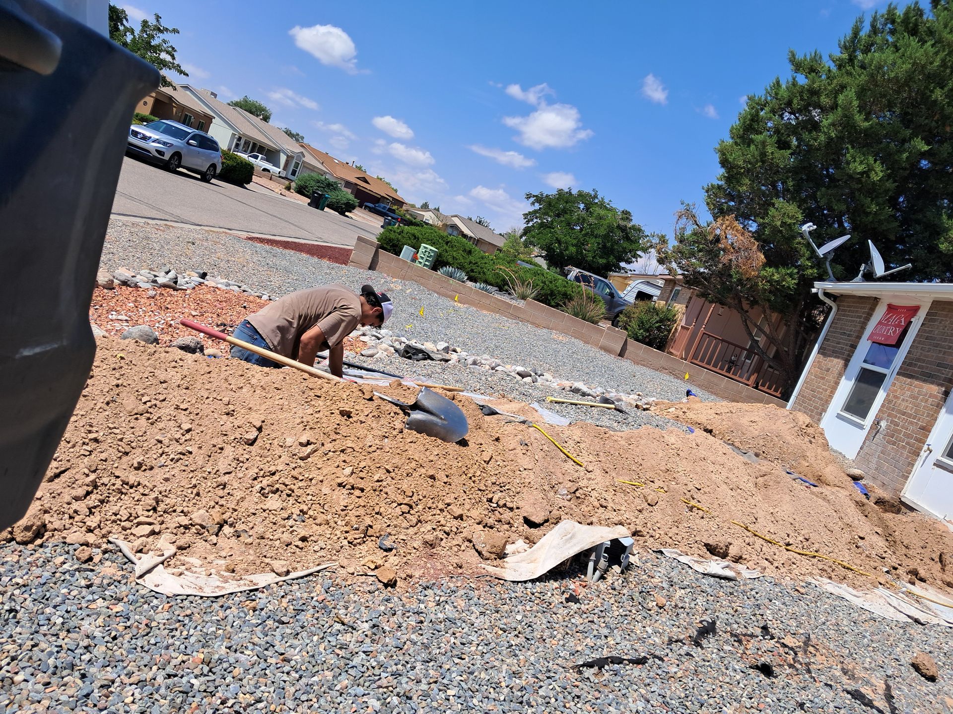 Person shoveling dirt on a sloped residential landscape; dirt pile, gravel, houses, blue sky.