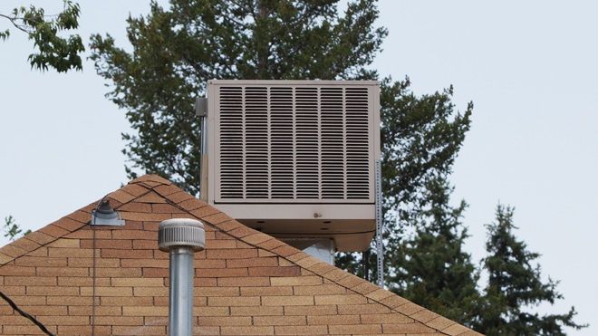 Rooftop air cooler on a brick house. Green trees in the background, gray sky.