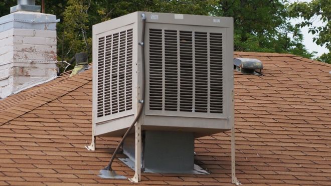 An evaporative cooler on a brown shingled roof next to a white chimney.