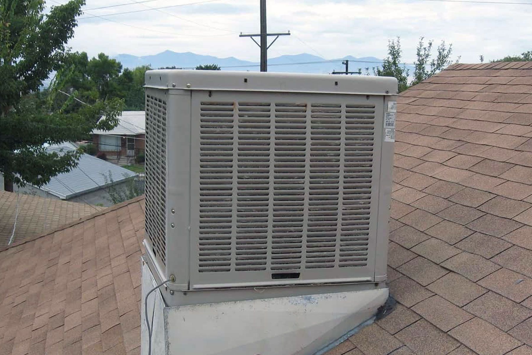Gray swamp cooler on a rooftop with brown shingles, overlooking a neighborhood and mountains.