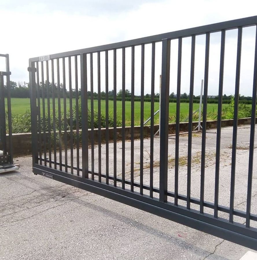 Un cancello scorrevole in metallo, verniciato di grigio scuro, poggiato su una superficie pavimentata con un campo aperto sullo sfondo.