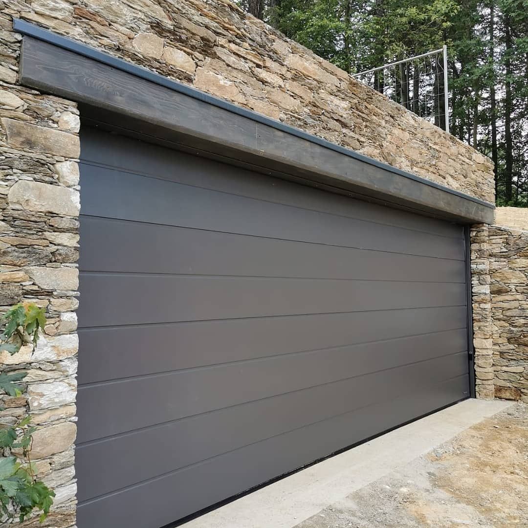 Una moderna porta da garage sezionale grigio scuro, incastonata in un muro di pietra rustica con vialetto d'accesso in cemento.