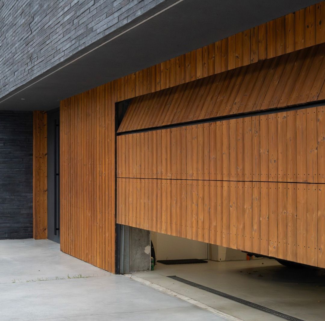 Una moderna porta da garage parzialmente aperta, caratterizzata da listelli verticali in legno che contrastano con una parete in pietra grigio scuro e un pavimento in cemento.