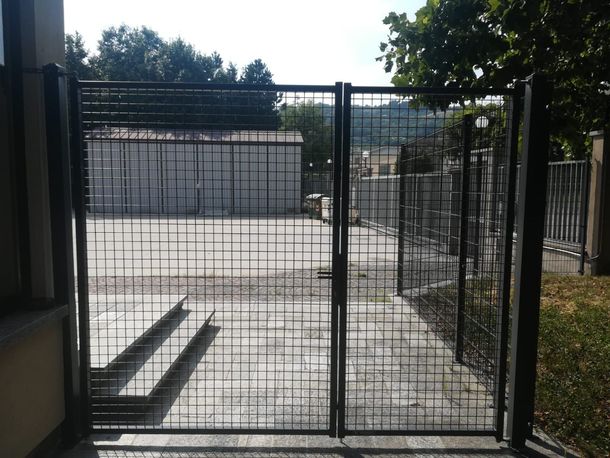 Un doppio cancello metallico con una griglia nera si erge chiuso davanti a un cortile pavimentato e a un piccolo edificio.