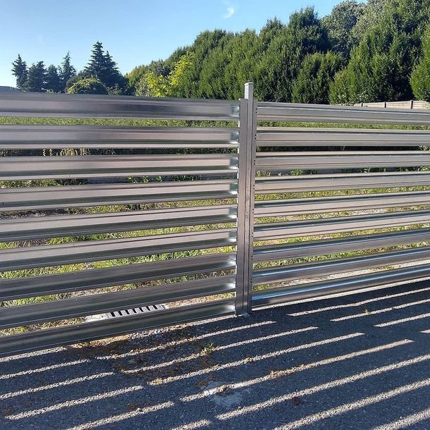 Una recinzione metallica con doghe orizzontali sullo sfondo di alberi verdi e un cielo azzurro limpido.