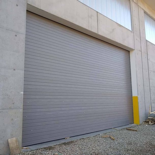 Una porta da garage avvolgibile in metallo grigio, chiusa e incastonata in un muro esterno di cemento, con un segnale di sicurezza giallo sul lato destro.