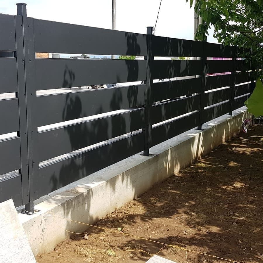 Una recinzione metallica nera con doghe orizzontali installata su una base di cemento in un cortile.