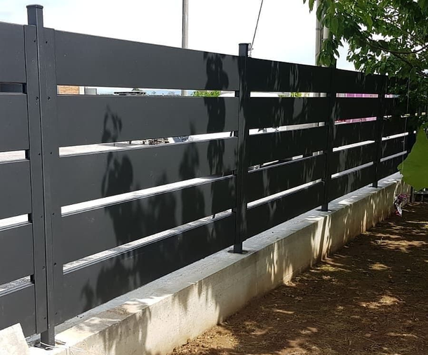 Una moderna recinzione a doghe orizzontali in metallo nero, montata su una base di cemento, vista da un'angolazione diversa in un ambiente esterno.