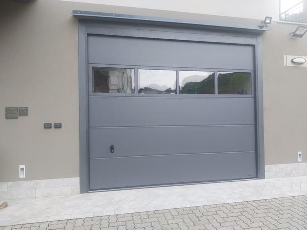 Una moderna porta da garage sezionale grigio scuro con una fila di quattro finestre orizzontali installata su una parete esterna di colore chiaro.