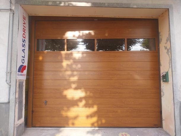 Una porta da garage sezionale con finitura effetto legno e una fila di finestre rettangolari nella parte superiore, installata in un telaio di cemento.