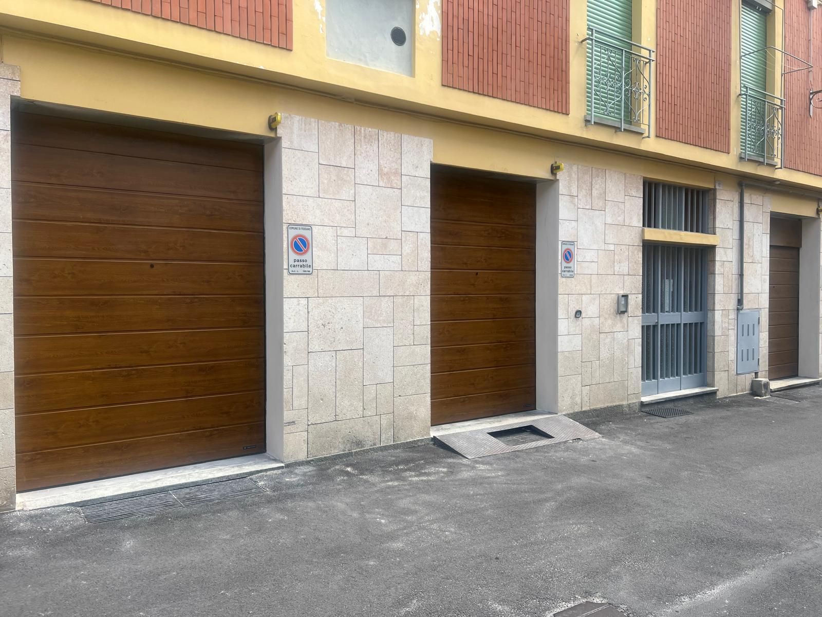 Vista a livello stradale di un edificio in pietra chiara con due portoni da garage marroni e un ingresso con pannelli in vetro.