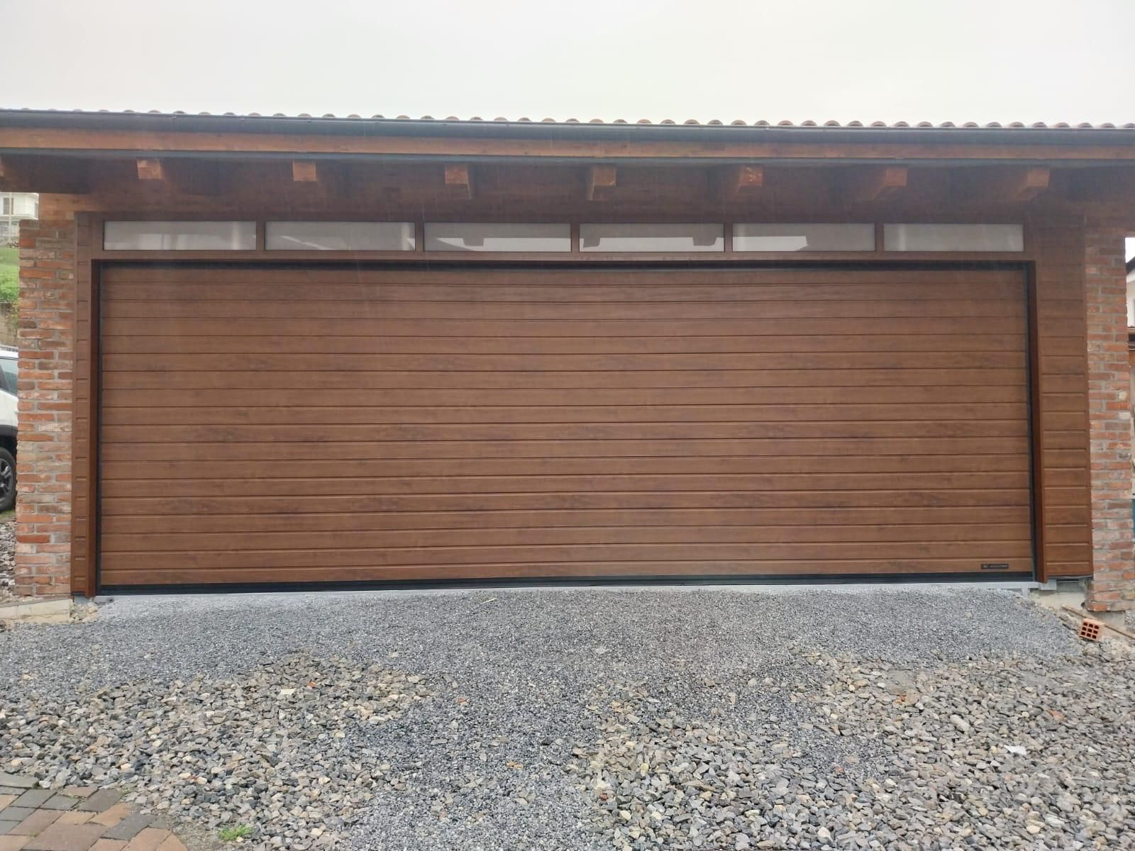 Una moderna porta da garage con pannelli in legno marrone e una fila di finestre a cleristorio, incastonata in un muro di pietra sopra un vialetto di ghiaia.