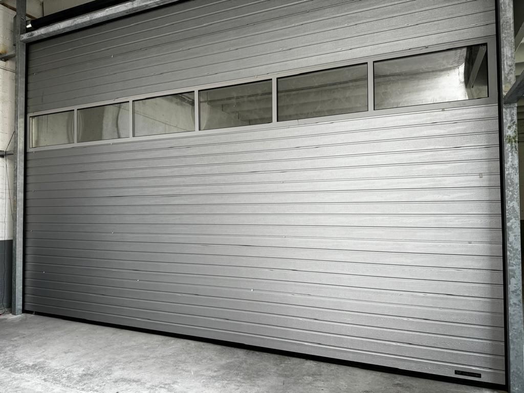 Una porta da garage chiusa in metallo con lamelle orizzontali, di colore grigio, con una fila di cinque finestre rettangolari nella parte superiore.