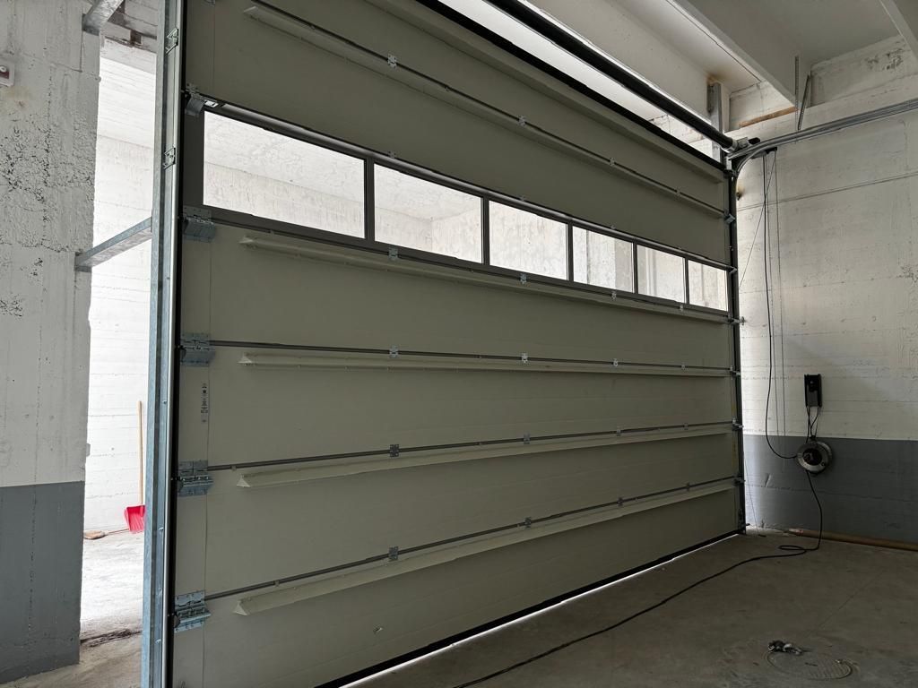 Una porta da garage sezionale industriale di colore chiaro con una fila di finestre rettangolari, installata all'interno di uno spazio in cemento.