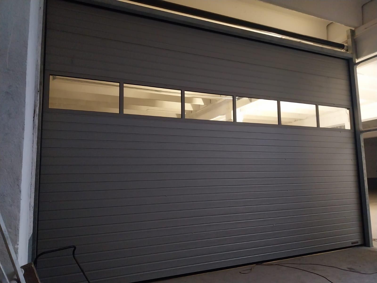 Una porta da garage sezionale grigia con una fila di finestre rettangolari nella parte superiore, installata su un edificio di colore chiaro.