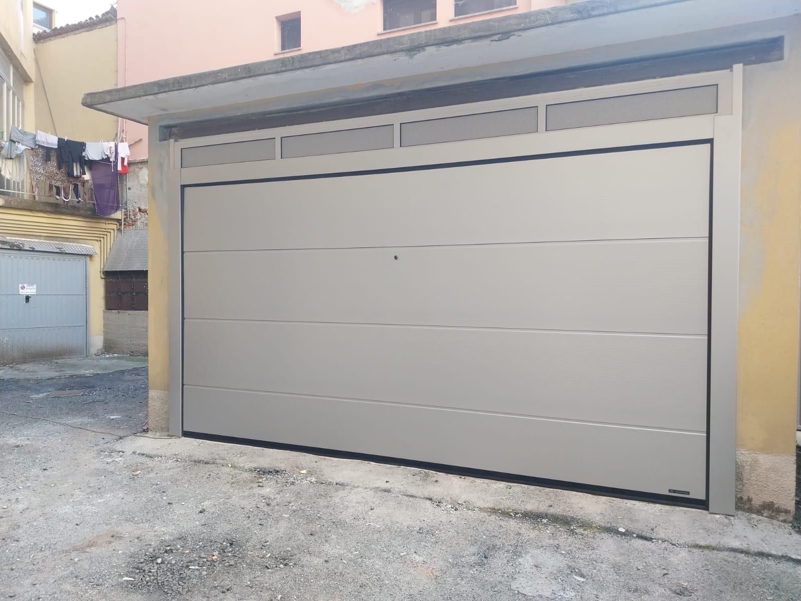 Una moderna porta da garage sezionale di colore grigio chiaro con pannelli orizzontali, installata su una facciata esterna di un edificio di colore giallo.