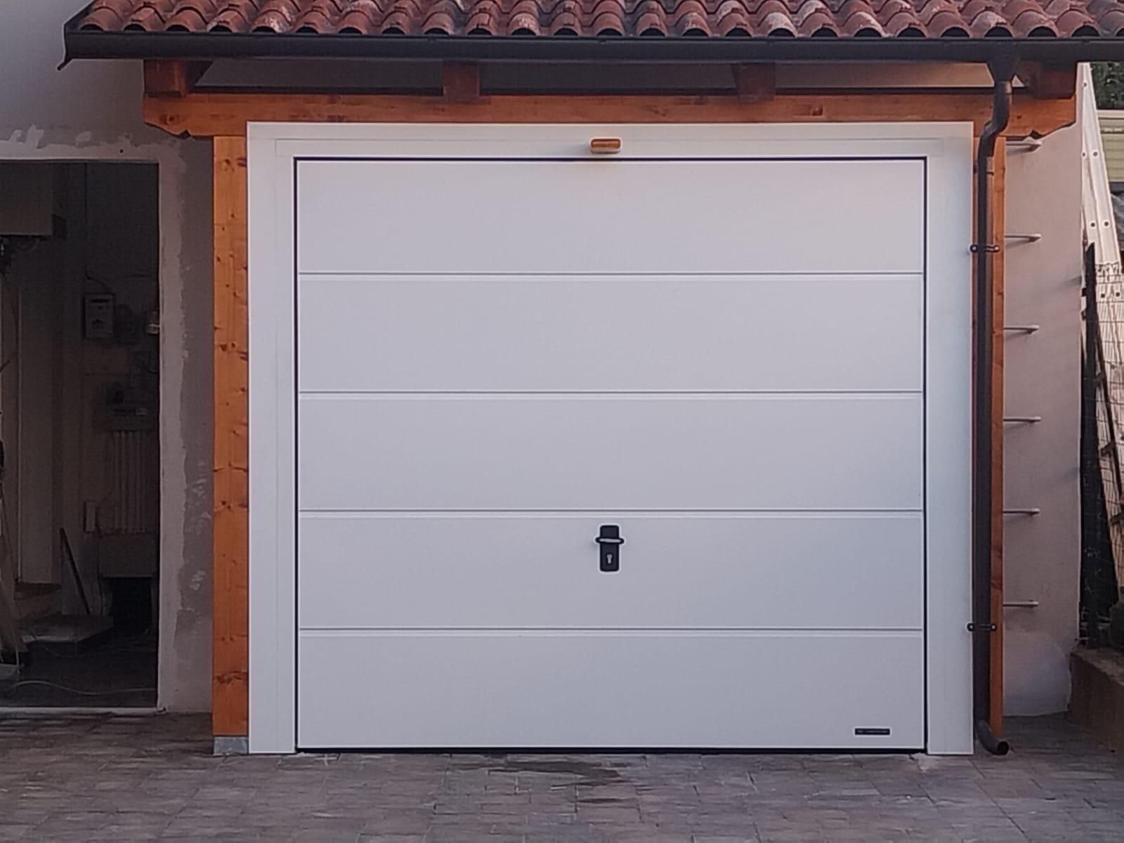 Una porta da garage bianca a pannelli orizzontali con serratura centralizzata, inserita in un telaio di legno sotto un tetto di tegole.