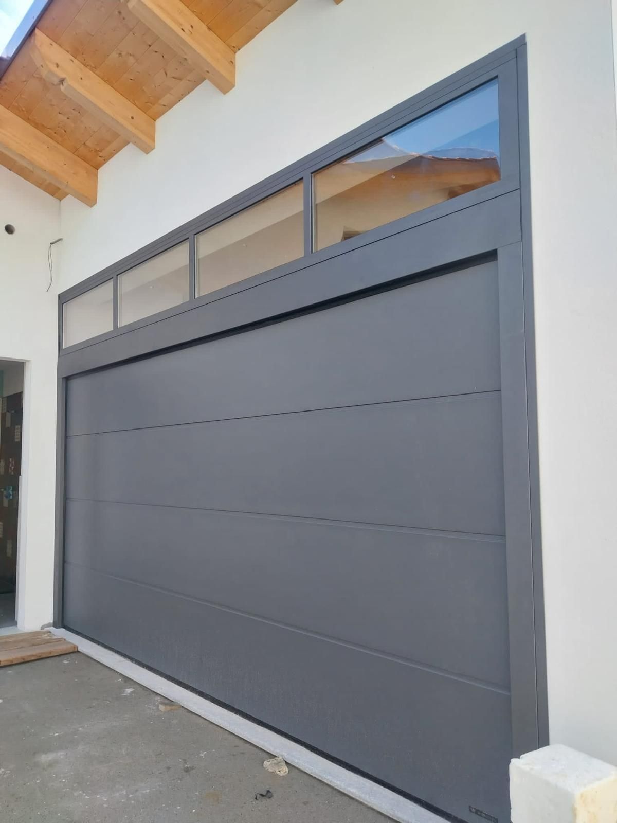 Una moderna porta da garage grigia con un design a pannelli orizzontali e una fila di finestre a lunetta in vetro, installata su una parete bianca.